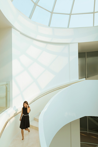 Young Chinese woman descending walking exploring exploration discovery admiration awe wonder curiosity staircase stairs marble banister design interior modern contemporary architectural architecture building with skylight window looking forward in admiration