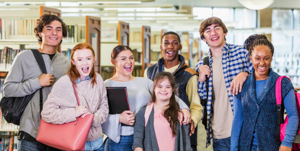 High school students in library, girl with down syndrome A multi-ethnic group of seven high school students, 15 to 17 years old, standing together in a library, smiling and looking at the camera. The girl in the middle has down syndrome. teenagers only stock pictures, royalty-free photos & images