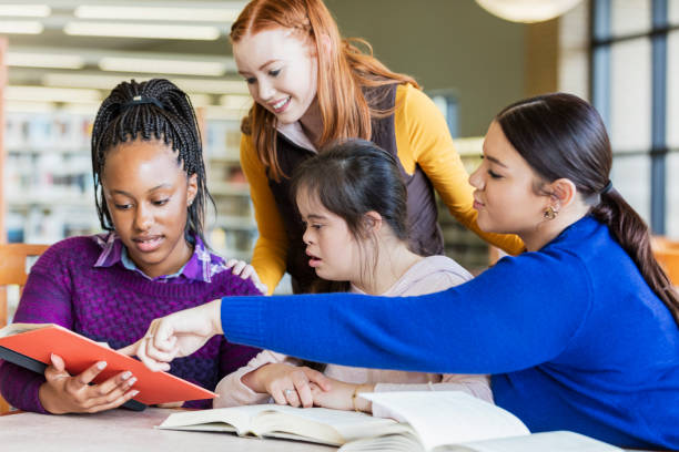 Teenage girl with down syndrome and friends studying A group of four multi-ethnic teenage girls at a table in the library, reading and studying together. The one sitting in the middle has down syndrome.  She is 17 years old mixed race Hispanic and Caucasian. She and her friends are looking at a book the 15 year old African-American girl is holding. teenagers only teenager multi ethnic group student stock pictures, royalty-free photos & images