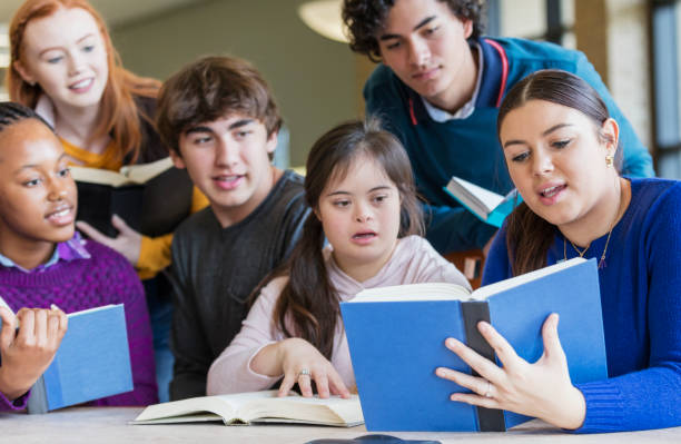 Teenage girl with down syndrome and friends reading A group of six multi-ethnic teenagers at a table in the library, reading and studying together. The one wearing the pink hooded shirt sitting in the middle has down syndrome. She is 17 years old mixed race Hispanic and Caucasian. The girl sitting next to her is holding up her book and they are all looking at it. teenagers only teenager multi ethnic group student stock pictures, royalty-free photos & images