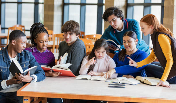 Teenage girl with down syndrome and friends studying A group of seven multi-ethnic teenagers at a table in the library, reading and studying together. The one sitting in the middle wearing the pink hooded shirt has down syndrome. She is 17 years old mixed race Hispanic and Caucasian. She and her friends, teenage boys and girls, 15 to 17 years old, are smiling, looking down at their books, conversing. teenagers only teenager multi ethnic group student stock pictures, royalty-free photos & images