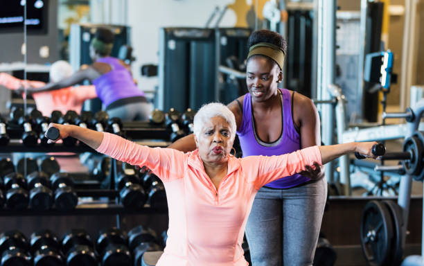 African-American fitness instructor helping senior woman A young African-American woman in her 20s working as a personal trainer or fitness instructor in a gym. She is helping a senior African-American woman in her 60s who grimacing as she lifts hand weights. fitness trainer stock pictures, royalty-free photos & images