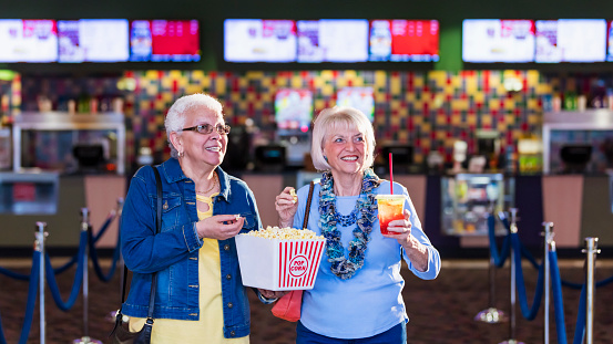 Two multi-ethnic senior women having fun together going to see a movie at the theater. They are carrying popcorn and drinks from the concession stand, smiling and walking. The Hispanic woman wearing eyeglasses is in her 60s and her friend is in her 70s.
