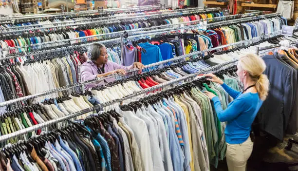 Two multi-ethnic women working in a clothing store. The Caucasian woman is in her 50s, and her coworker is a senior African-American woman in her 60s. They are talking to each other over racks of men's shirts.