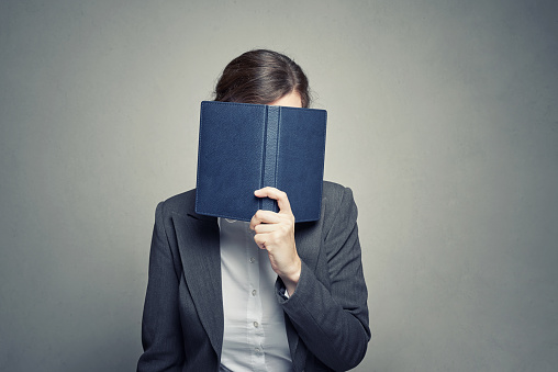 business woman in white blouse and suit cover her face with organizer diary on gray background