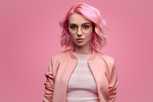 Positive young woman in stylish glasses looking at camera while standing on pink background
