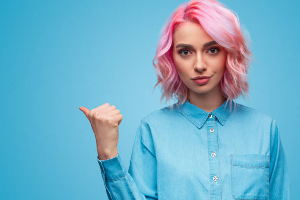 Woman with pink hair pointing aside Lovely young lady with pink hair looking at camera and pointing aside with thumb while standing on blue background smirking stock pictures, royalty-free photos & images