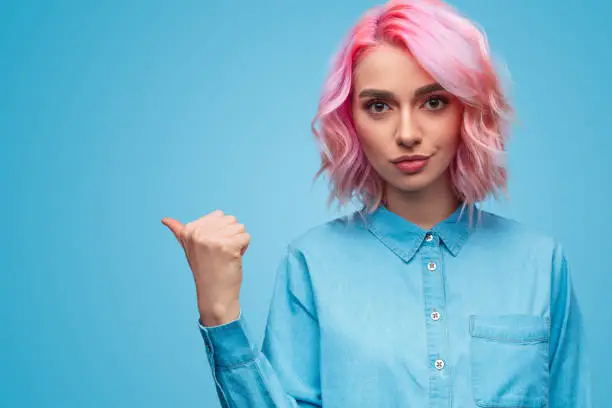 Lovely young lady with pink hair looking at camera and pointing aside with thumb while standing on blue background
