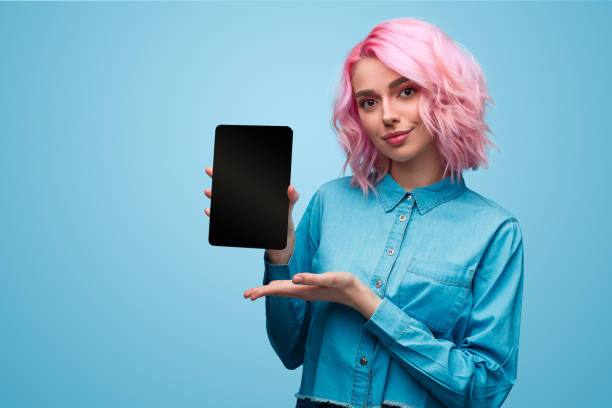 Charming young millennial woman showing tablet Pretty informal woman with pink hair wearing blue shirt and demonstrating new tablet on blue background pink hair stock pictures, royalty-free photos & images