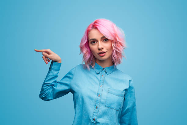 Crazy young woman pointing aside Funny young lady with pink hair looking at camera with crazy face expression and pointing aside while standing on blue background grimacing stock pictures, royalty-free photos & images
