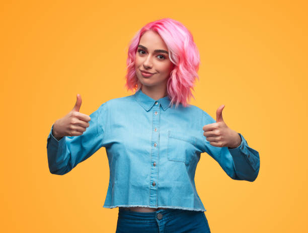 Lady with pink hair gesturing thumb-ups Pretty young female with pink hair smiling and looking at camera while standing on yellow background and showing thumb-up gesture with both hands pink promise stock pictures, royalty-free photos & images