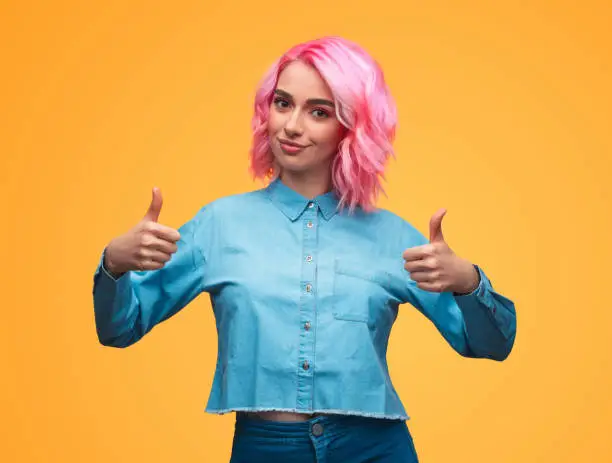 Pretty young female with pink hair smiling and looking at camera while standing on yellow background and showing thumb-up gesture with both hands