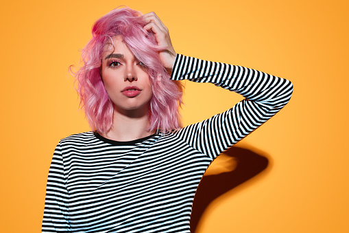 Beautiful young lady in striped outfit rumpling pink hair and looking at camera while standing on yellow background