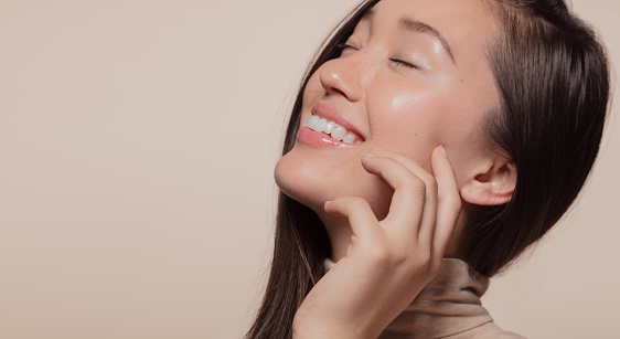 Close up of beautiful asian woman with hand on cheeks over beige background with copy space on one side. Charming korean female face with beautiful skin.