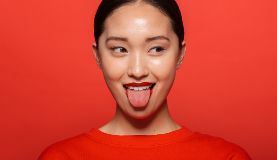 Close up of young asian woman sticking out tongue and looking away. Korean female model making funny face against red background.