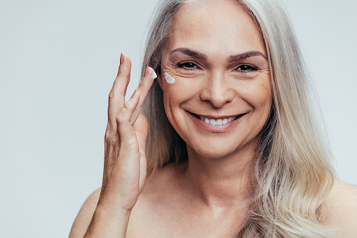 Smiling mid adult caucasian woman applying anti aging cream on her face. Senior female woman applying moisturizer on her face against grey background.