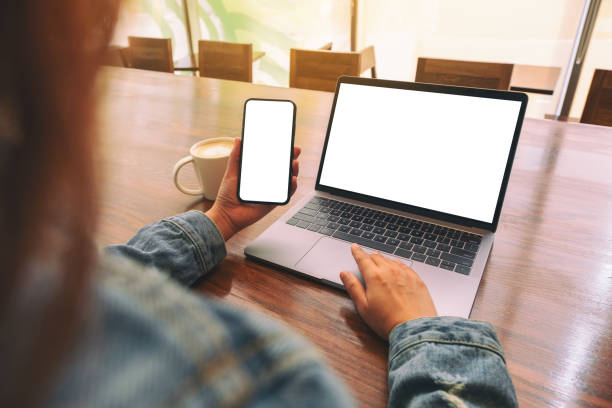 istock Mockup image of a woman touching on laptop touchpad with blank white desktop screen while using mobile phone 1140958263