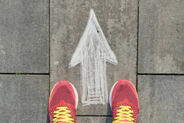 Arrow sign painted on gray sidewalk with women legs in sneakers, top view.