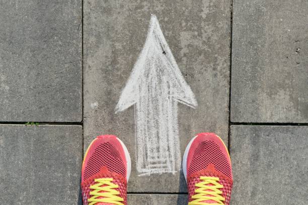 стрелка знак нарисован на сером тротуаре с женскими ногами в кроссовках, вид сверху - choice street road beginnings стоковые фото и изоб�ражения