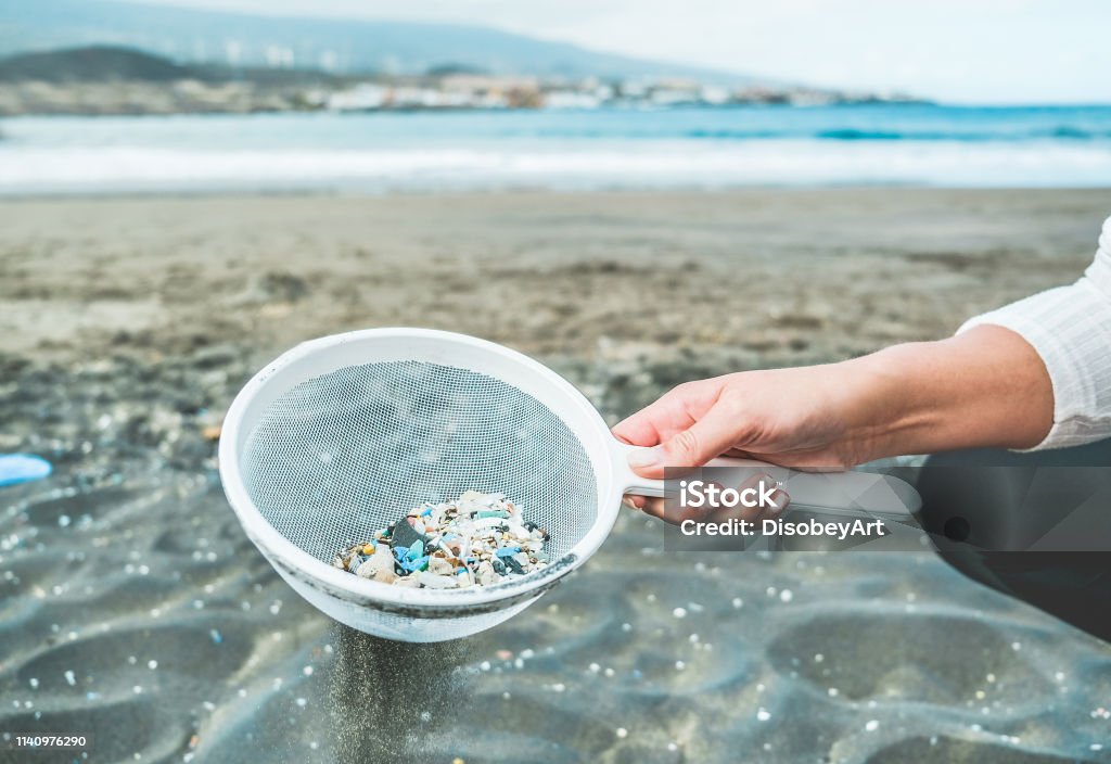 Giovane donna che pulisce le microplastiche dalla sabbia sulla spiaggia - Problema ambientale, inquinamento e concetto di avvertimento dell'ecolosistema - Focus sulla mano - Foto stock royalty-free di Microplastica