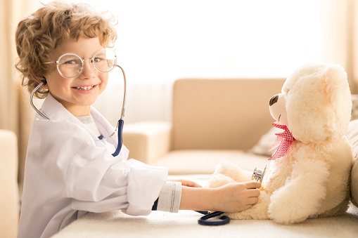 Cute happy little boy in whitecoat and eyeglasses looking at you while playing doctor with teddybear