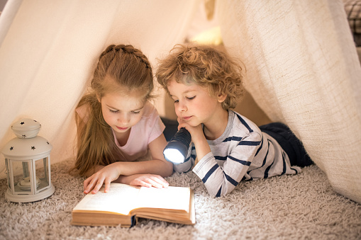 Two cuties in casualwear lying inside selfmade tent on the floor and using lantern while reading book of tales
