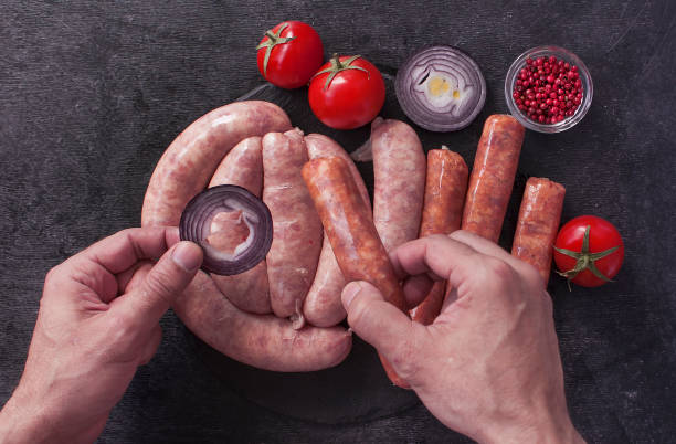 Men's hands prepare an assortment of fresh raw sausages with tomatoes, onions and spices on a black wooden chopping Board. The view from the top.