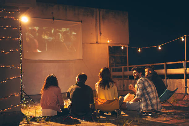 Friends watching a movie on a building rooftop terrace Group of young friends watching a movie on a building rooftop terrace, eating popcorn, drinking beer and having fun movie night friends stock pictures, royalty-free photos & images