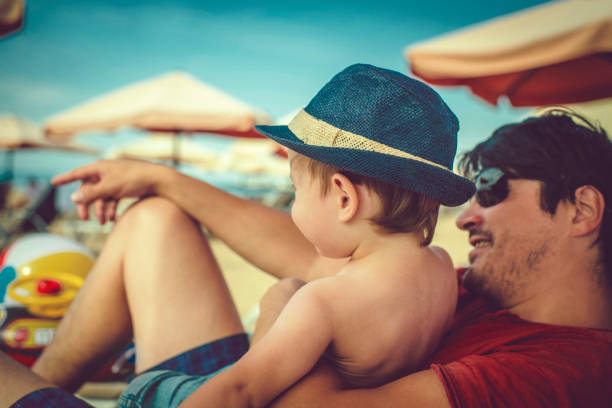 A father playfully lifts his young son into the air at the beach. The boy, holding an orange shovel, laughs with delight as they enjoy their time by the sea.