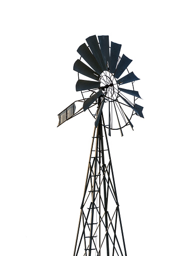 Low angle view of an old-fashioned, multi-bladed, metal wind pump atop a lattice tower in backlight, cut out on white background.