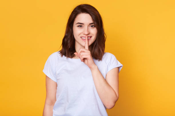 conceito da língua do corpo. a fêmea nova de sorriso bonita com expressão feliz, desgasta a camisa de t ocasional, demonstra o gesto do silêncio, mantem o dedo dianteiro nos bordos, isolados no fundo amarelo do estúdio. - indexfinger - fotografias e filmes do acervo