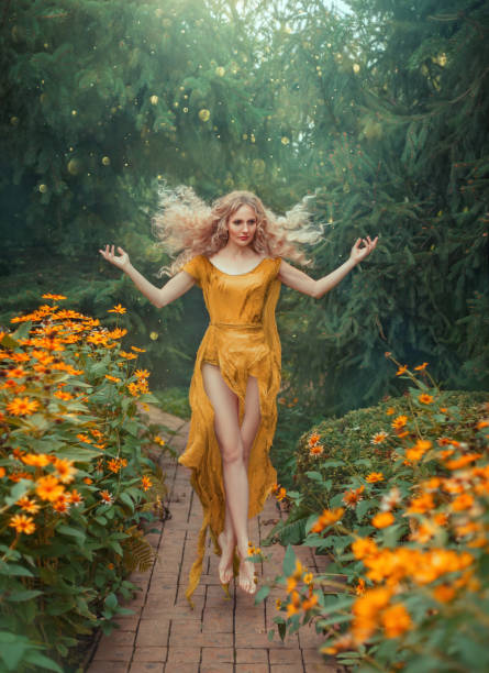 fairy atrativo misterioso da flor no vestido amarelo claro com trem longo e os pés abertos no salto na floresta com flores brilhantes. senhora com cabelo louro que voa na luz solar brilhante, arte que processa a foto - fairy forest women magic - fotografias e filmes do acervo