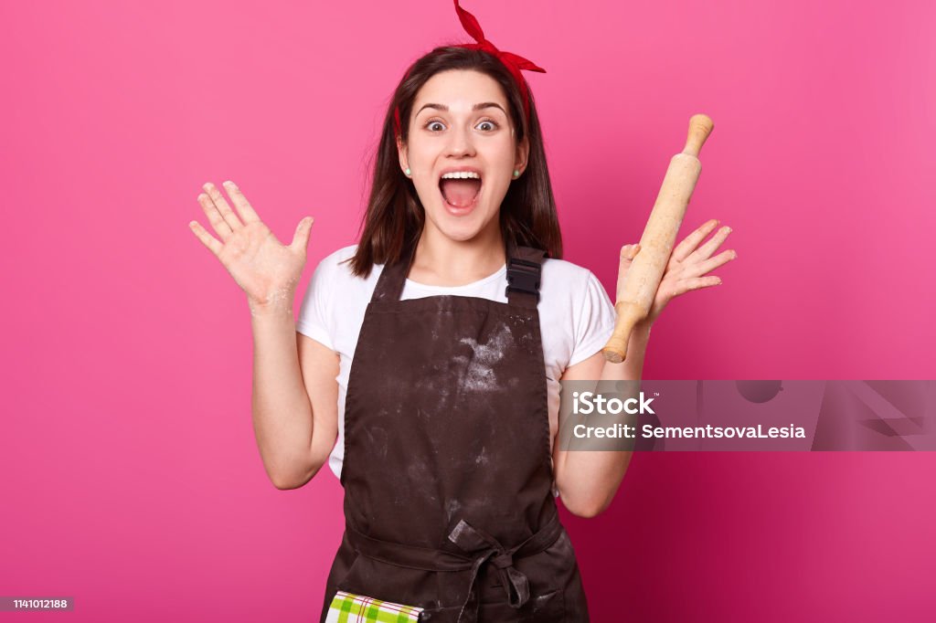 Baker young woman puts her hands up holding rolling pin, wears brown apron, white tshirt, opens her mouth widely. Adorable cute female is in high spirits while cooking new dishes. Cook concept. Adult Stock Photo