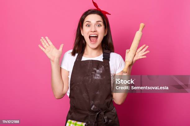 Baker Young Woman Puts Her Hands Up Holding Rolling Pin Wears Brown Apron White Tshirt Opens Her Mouth Widely Adorable Cute Female Is In High Spirits While Cooking New Dishes Cook Concept Stock Photo - Download Image Now
