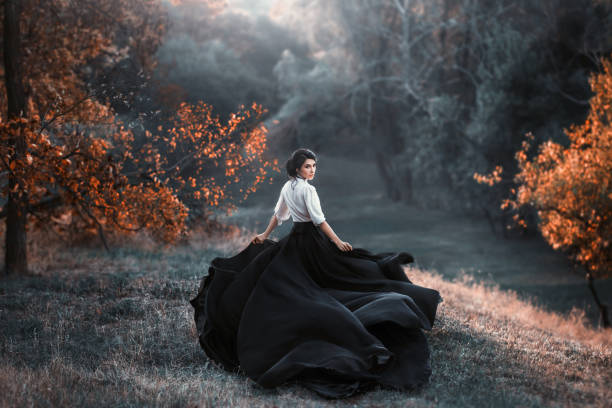 mysterious girl with dark hair and a long flying black vintage dress runs through the woods, a charming ambush, a great attractive witch turns into a crow, waving a train like bird wings - witch beauty beautiful women imagens e fotografias de stock