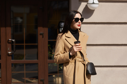 Beautiful brunette young woman holds cup of coffee in her hands, walks along sunlighted streets, wears long fashionable mustard coat, has black accessories, bright makeup. Street fashion concept.
