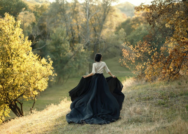 princesse dans une robe vintage. marchez le long des collines pittoresques d’automne au coucher du soleil. un long train de jupe noire flottaient sur la course. photo d’une fille brune avec un dos sans visage. photo artistique - color image halloween people elegance photos et images de collection