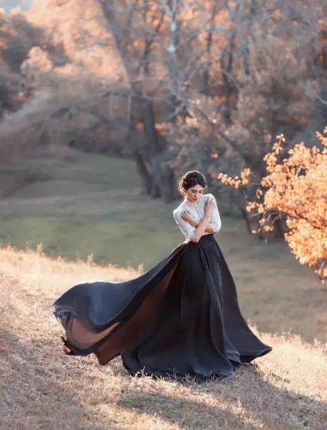 The lady in a vintage dress strolls along the picturesque, golden, autumn hills at sunset. A long train of black skirt fluttering in the wind. The girl is hugging herself from the cold. Artistic Photo
