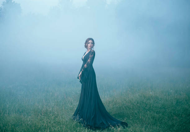 una ragazza in un abito lungo nero che cammina lungo ia una radura nella fitta nebbia. spaventato, bello, strega ia andare nella foresta, alla ricerca di una casa. foto d'arte. costume di halloween. modello. oscuro. mistico. cattivo - women gothic style sensuality mystery foto e immagini stock