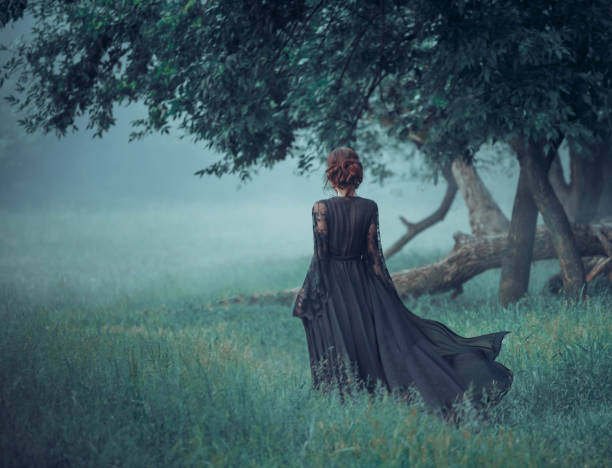 ragazza con i capelli rossi che camminano dalla foresta scura, indossando un lungo vestito nero con rimorchio che ondeggia nel vento. strega. foto d'arte in colori caldi, alberi di smeraldo, erba, fiaba, halloween - women gothic style sensuality mystery foto e immagini stock