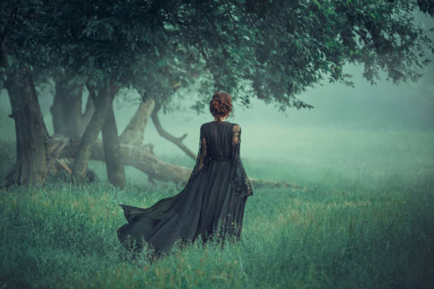 赤い髪の少女は、暗い森から歩いて、風の中で手を振っているトレーラーと長い黒いドレスを着ています。魔女。暖かい色、エメラルドの木、草、おとぎ話、ハロウィーンのアート写真 - women gothic style sensuality mystery ストックフォトと画像