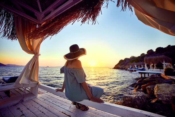 Woman in hat relaxing by the sea in a luxurious beachfront hotel resort at sunset enjoying perfect beach holiday vacation in Bodrum, Turkey. Outdoors Seascape Summer Travel Concept mujer en el sombrero de relax junto al mar en un lujoso complejo hotelero frente a la playa al atardecer disfrutando de perfectas vacaciones de vacaciones en las playas en bodrum, turquía. el concepto de viaje de verano en exterior seascape