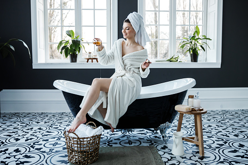 A cute young woman wearing a white robe and having a towel on her head spend time in the bathroom. Leisure