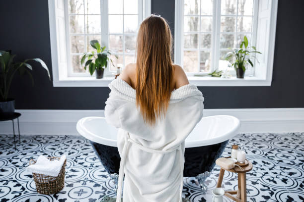 istock Beautiful light bathroom with large windows. A young woman removes her white robe while standing at the bath 1141036123