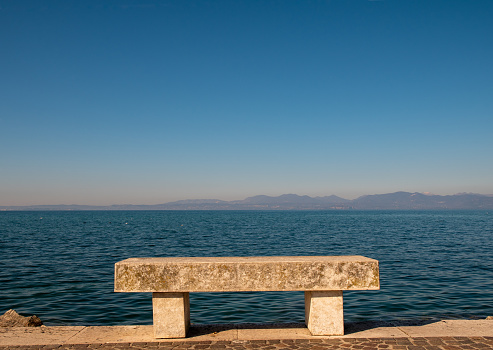 A stone bench on the lakefront of Lazise with Lake Garda and the coastline in the background, Veneto, Italy