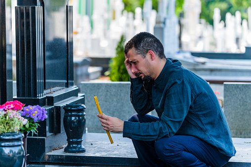 Mid Aged Tired Man is Sitting Near a Grave in the Cemetery and Mourning Over the Deceased. Widower in Black Clothes Kneeling in Front of the Headstone, Holding a Candle and Grieve for Family Loss.