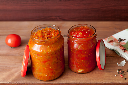 Homemade prepared two fermented vegetable glass jars with carrot, red tomatoes and salsa with spice on rustic wooden background. Vegetarian food concept
