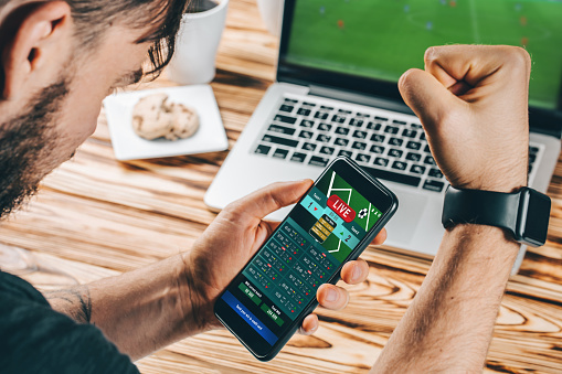 Lucky man celebrating victory after making bets using gambling mobile application on his phone. Football match online broadcast on laptop screen on the background.