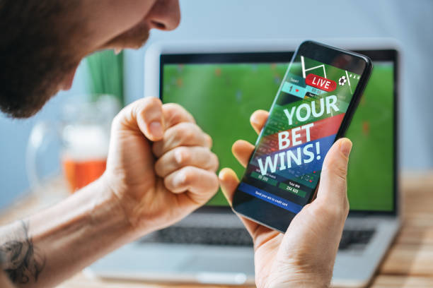 Man celebrating victory after making bets at bookmaker website Lucky man celebrating victory after making bets using gambling mobile application on his phone. Football match online broadcast on laptop screen on the background. number one sports betting site stock pictures, royalty-free photos & images