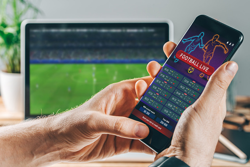 Close up cropped shot of male hands making bets using gambling mobile application on his phone. Man watching football match online broadcast on his laptop waiting for winning results.